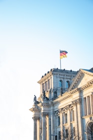 A historic architectural building with classical columns and statues. At the top, a German flag waves against a clear blue sky.