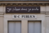A building facade displays two signs. The top sign, in a playful cursive font, reads 'Je pisse donc je suis,' while the bottom sign in bold letters states 'W-C Publics.' The exterior is a beige stone with visible architectural details, such as molding and window frames.