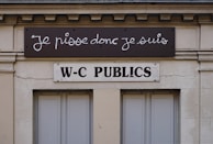A building facade displays two signs. The top sign, in a playful cursive font, reads 'Je pisse donc je suis,' while the bottom sign in bold letters states 'W-C Publics.' The exterior is a beige stone with visible architectural details, such as molding and window frames.