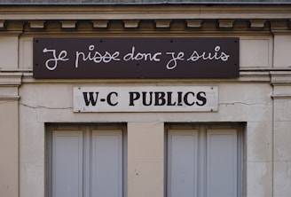 A building facade displays two signs. The top sign, in a playful cursive font, reads 'Je pisse donc je suis,' while the bottom sign in bold letters states 'W-C Publics.' The exterior is a beige stone with visible architectural details, such as molding and window frames.