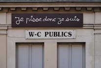 A building facade displays two signs. The top sign, in a playful cursive font, reads 'Je pisse donc je suis,' while the bottom sign in bold letters states 'W-C Publics.' The exterior is a beige stone with visible architectural details, such as molding and window frames.