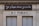 A building facade displays two signs. The top sign, in a playful cursive font, reads 'Je pisse donc je suis,' while the bottom sign in bold letters states 'W-C Publics.' The exterior is a beige stone with visible architectural details, such as molding and window frames.
