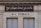 A building facade displays two signs. The top sign, in a playful cursive font, reads 'Je pisse donc je suis,' while the bottom sign in bold letters states 'W-C Publics.' The exterior is a beige stone with visible architectural details, such as molding and window frames.