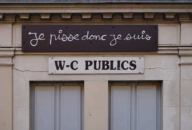 A building facade displays two signs. The top sign, in a playful cursive font, reads 'Je pisse donc je suis,' while the bottom sign in bold letters states 'W-C Publics.' The exterior is a beige stone with visible architectural details, such as molding and window frames.