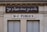 A building facade displays two signs. The top sign, in a playful cursive font, reads 'Je pisse donc je suis,' while the bottom sign in bold letters states 'W-C Publics.' The exterior is a beige stone with visible architectural details, such as molding and window frames.