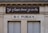 A building facade displays two signs. The top sign, in a playful cursive font, reads 'Je pisse donc je suis,' while the bottom sign in bold letters states 'W-C Publics.' The exterior is a beige stone with visible architectural details, such as molding and window frames.