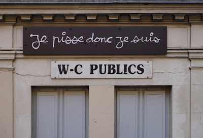 A building facade displays two signs. The top sign, in a playful cursive font, reads 'Je pisse donc je suis,' while the bottom sign in bold letters states 'W-C Publics.' The exterior is a beige stone with visible architectural details, such as molding and window frames.
