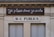 A building facade displays two signs. The top sign, in a playful cursive font, reads 'Je pisse donc je suis,' while the bottom sign in bold letters states 'W-C Publics.' The exterior is a beige stone with visible architectural details, such as molding and window frames.