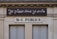 A building facade displays two signs. The top sign, in a playful cursive font, reads 'Je pisse donc je suis,' while the bottom sign in bold letters states 'W-C Publics.' The exterior is a beige stone with visible architectural details, such as molding and window frames.