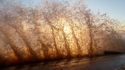 Water splashes dramatically against a surface, capturing sunlight that causes the droplets to glow with warm hues. The scene creates an interplay of light and water, resulting in a dynamic and visually striking pattern.