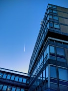 A modern office building with a grid of reflective windows stretching upwards towards the clear blue sky. A trail from an aircraft is visible, appearing as a thin, white streak in the sky, suggesting simplicity and minimalism in the urban landscape.