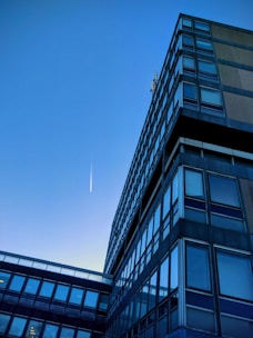 A modern office building with a grid of reflective windows stretching upwards towards the clear blue sky. A trail from an aircraft is visible, appearing as a thin, white streak in the sky, suggesting simplicity and minimalism in the urban landscape.