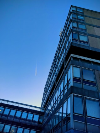 A modern office building with a grid of reflective windows stretching upwards towards the clear blue sky. A trail from an aircraft is visible, appearing as a thin, white streak in the sky, suggesting simplicity and minimalism in the urban landscape.