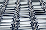 A repeating pattern featuring rows of metallic conveyor rollers arranged horizontally, creating a sense of industrial symmetry. The rollers have a shiny, reflective surface and are uniformly aligned, suggesting movement or transportation capabilities.