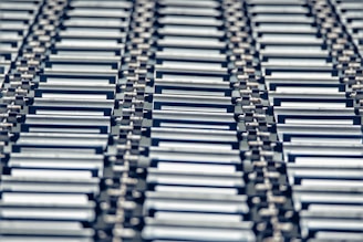 A repeating pattern featuring rows of metallic conveyor rollers arranged horizontally, creating a sense of industrial symmetry. The rollers have a shiny, reflective surface and are uniformly aligned, suggesting movement or transportation capabilities.