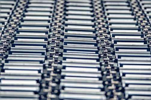 A repeating pattern featuring rows of metallic conveyor rollers arranged horizontally, creating a sense of industrial symmetry. The rollers have a shiny, reflective surface and are uniformly aligned, suggesting movement or transportation capabilities.