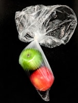 A green apple and a red apple enclosed in a transparent plastic bag against a black background. The bag is loosely tied at the top, crumpled, and shiny, reflecting light.