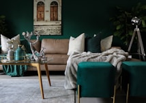 A chic and cozy living room features a beige sofa adorned with plush white and green pillows. In front of the sofa, a glass coffee table with golden legs displays a decorative tray with vases and a silver sculpture. The background includes a large, leafy green plant and a vintage-style floor lamp with a tripod stand. The walls are painted a deep green, complementing the teal ottomans at the forefront, while a piece of art depicting an old building with wooden doors hangs above the couch. A soft, textured gray blanket is draped over the furniture, enhancing the inviting atmosphere.