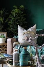 A cozy and stylish living room setting with a focus on earthy and modern decor elements. The scene includes a lush, leafy plant in the background and a comfortable beige sofa adorned with a white pillow featuring gold patterns. In the foreground, there is a decorative tray on a glass table, holding a green ceramic bottle, an hourglass, and a geometric ornament. The room is accentuated with rich green tones and natural textures, creating a calming and elegant atmosphere.