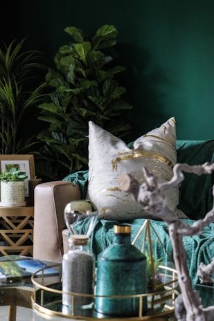 A cozy and stylish living room setting with a focus on earthy and modern decor elements. The scene includes a lush, leafy plant in the background and a comfortable beige sofa adorned with a white pillow featuring gold patterns. In the foreground, there is a decorative tray on a glass table, holding a green ceramic bottle, an hourglass, and a geometric ornament. The room is accentuated with rich green tones and natural textures, creating a calming and elegant atmosphere.