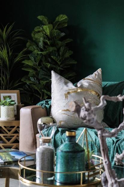 A cozy and stylish living room setting with a focus on earthy and modern decor elements. The scene includes a lush, leafy plant in the background and a comfortable beige sofa adorned with a white pillow featuring gold patterns. In the foreground, there is a decorative tray on a glass table, holding a green ceramic bottle, an hourglass, and a geometric ornament. The room is accentuated with rich green tones and natural textures, creating a calming and elegant atmosphere.