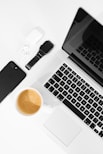 A well-arranged flat lay of modern tech gadgets placed on a white surface, including a sleek black smartphone, wireless earbuds in a white case, a black smartwatch, a silver laptop with a black keyboard, and a cup of coffee with a rich crema.