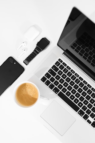 A well-arranged flat lay of modern tech gadgets placed on a white surface, including a sleek black smartphone, wireless earbuds in a white case, a black smartwatch, a silver laptop with a black keyboard, and a cup of coffee with a rich crema.