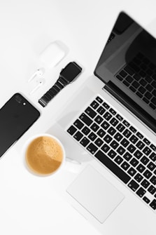 A well-arranged flat lay of modern tech gadgets placed on a white surface, including a sleek black smartphone, wireless earbuds in a white case, a black smartwatch, a silver laptop with a black keyboard, and a cup of coffee with a rich crema.