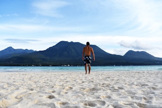man walking to the water with the distance of mountain during daytime