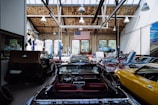 A spacious automotive workshop with a high wooden ceiling and large arched windows. Several classic cars in various colors, including yellow, black, and red, are parked with their hoods open. A large American flag hangs from the ceiling, and there's a vehicle lift with a blue car raised. The workshop has a clean, organized appearance with tools and equipment neatly arranged.