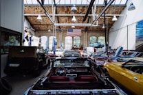 A spacious automotive workshop with a high wooden ceiling and large arched windows. Several classic cars in various colors, including yellow, black, and red, are parked with their hoods open. A large American flag hangs from the ceiling, and there's a vehicle lift with a blue car raised. The workshop has a clean, organized appearance with tools and equipment neatly arranged.