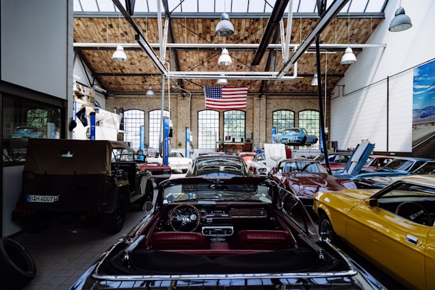 A spacious automotive workshop with a high wooden ceiling and large arched windows. Several classic cars in various colors, including yellow, black, and red, are parked with their hoods open. A large American flag hangs from the ceiling, and there's a vehicle lift with a blue car raised. The workshop has a clean, organized appearance with tools and equipment neatly arranged.