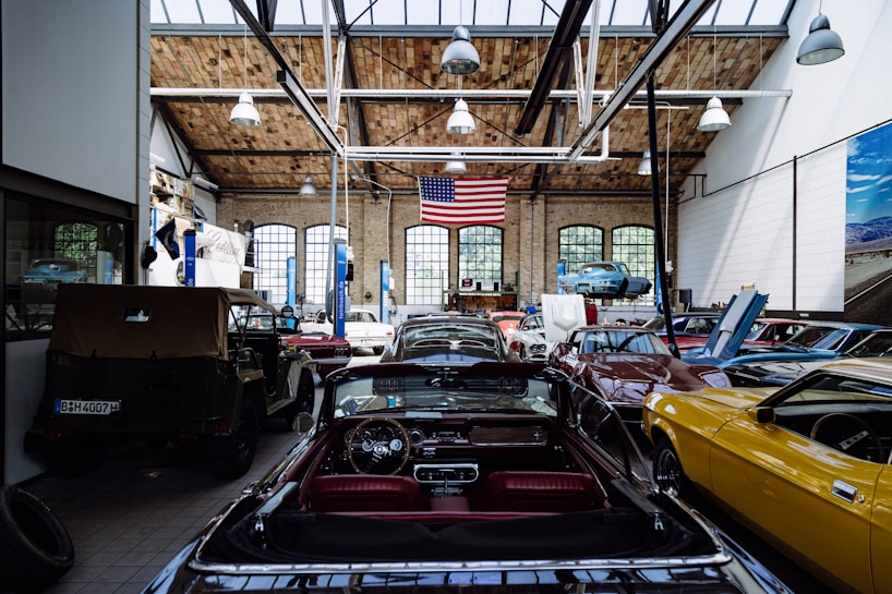 A spacious automotive workshop with a high wooden ceiling and large arched windows. Several classic cars in various colors, including yellow, black, and red, are parked with their hoods open. A large American flag hangs from the ceiling, and there's a vehicle lift with a blue car raised. The workshop has a clean, organized appearance with tools and equipment neatly arranged.