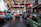 A spacious automotive workshop with several vintage and classic cars, some on lifts. The interior has a high ceiling with exposed beams and large windows allowing natural light. The walls are adorned with automotive posters and signs, including various brands. An organized yet busy atmosphere with tools and equipment visible.