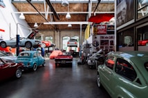 A spacious automotive workshop with several vintage and classic cars, some on lifts. The interior has a high ceiling with exposed beams and large windows allowing natural light. The walls are adorned with automotive posters and signs, including various brands. An organized yet busy atmosphere with tools and equipment visible.