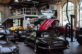 A vintage car garage with multiple classic cars, including some with convertible tops. One car is on a lift with its hood open, suggesting maintenance or repair. The setting is industrial, with high ceilings, large windows, and a metal mezzanine, filled with automotive equipment and tools.