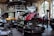 A vintage car garage with multiple classic cars, including some with convertible tops. One car is on a lift with its hood open, suggesting maintenance or repair. The setting is industrial, with high ceilings, large windows, and a metal mezzanine, filled with automotive equipment and tools.