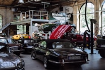 A vintage car garage with multiple classic cars, including some with convertible tops. One car is on a lift with its hood open, suggesting maintenance or repair. The setting is industrial, with high ceilings, large windows, and a metal mezzanine, filled with automotive equipment and tools.