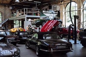 A vintage car garage with multiple classic cars, including some with convertible tops. One car is on a lift with its hood open, suggesting maintenance or repair. The setting is industrial, with high ceilings, large windows, and a metal mezzanine, filled with automotive equipment and tools.