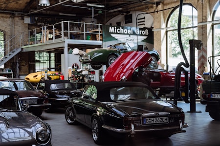 A vintage car garage with multiple classic cars, including some with convertible tops. One car is on a lift with its hood open, suggesting maintenance or repair. The setting is industrial, with high ceilings, large windows, and a metal mezzanine, filled with automotive equipment and tools.