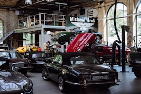 A vintage car garage with multiple classic cars, including some with convertible tops. One car is on a lift with its hood open, suggesting maintenance or repair. The setting is industrial, with high ceilings, large windows, and a metal mezzanine, filled with automotive equipment and tools.