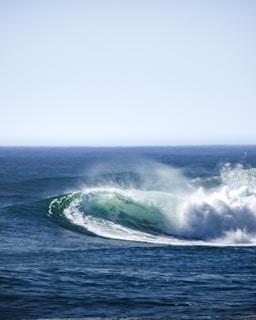 A powerful ocean wave curls and crashes, sending a mist of sea spray into the air. The wave is vibrant with shades of blue and green, against a backdrop of the open sea and a clear sky.