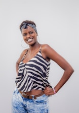 A person is standing with a confident and joyful expression, dressed in a zebra-striped sleeveless top and light blue jeans. A patterned headband is worn and large hoop earrings enhance the casual, fashionable style.