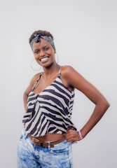 A person is standing with a confident and joyful expression, dressed in a zebra-striped sleeveless top and light blue jeans. A patterned headband is worn and large hoop earrings enhance the casual, fashionable style.