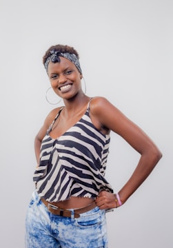 A person is standing with a confident and joyful expression, dressed in a zebra-striped sleeveless top and light blue jeans. A patterned headband is worn and large hoop earrings enhance the casual, fashionable style.