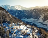 A picturesque winter landscape featuring snow-covered mountains and a serene valley. Cozy wooden chalets are scattered on a forested slope, surrounded by snow-laden trees. The sun casts a warm glow, highlighting the beauty of the landscape.