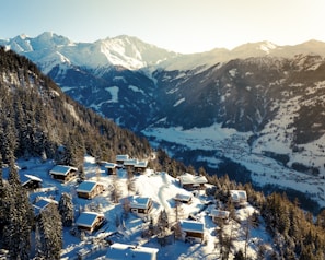 A picturesque winter landscape featuring snow-covered mountains and a serene valley. Cozy wooden chalets are scattered on a forested slope, surrounded by snow-laden trees. The sun casts a warm glow, highlighting the beauty of the landscape.