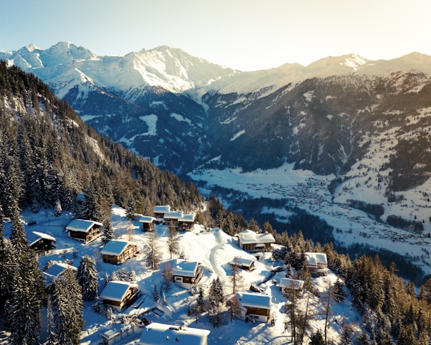 A picturesque winter landscape featuring snow-covered mountains and a serene valley. Cozy wooden chalets are scattered on a forested slope, surrounded by snow-laden trees. The sun casts a warm glow, highlighting the beauty of the landscape.