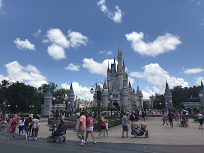 A large crowd of people is gathered in front of a fairy-tale castle with multiple spires. The sky is bright blue with scattered white clouds. Visitors are walking around, some pushing strollers, others taking photos. The scene is lively and energetic, typical of a theme park environment.