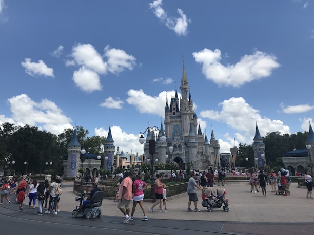 A large crowd of people is gathered in front of a fairy-tale castle with multiple spires. The sky is bright blue with scattered white clouds. Visitors are walking around, some pushing strollers, others taking photos. The scene is lively and energetic, typical of a theme park environment.